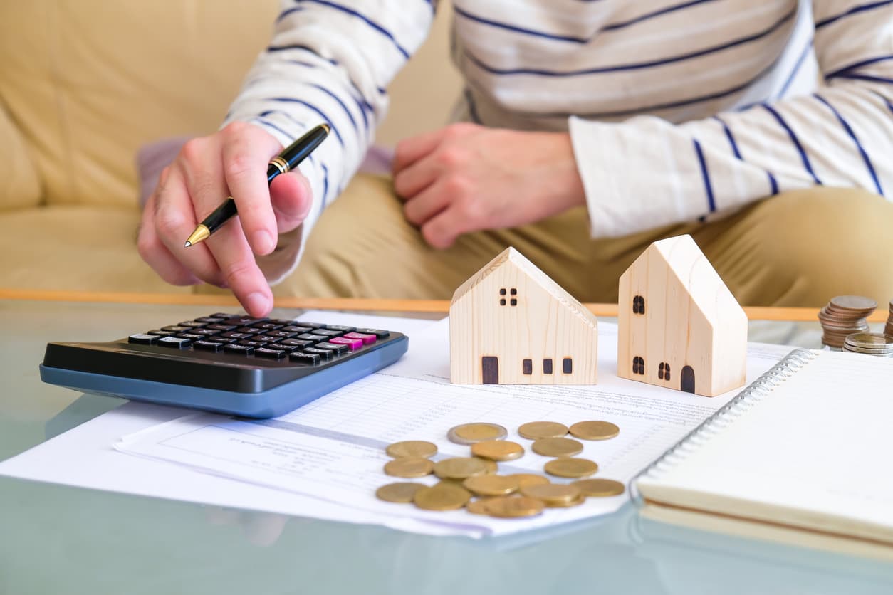 Man calculating sales compensation with a calculator, two wooden houses, and some pennies on a table in front of him