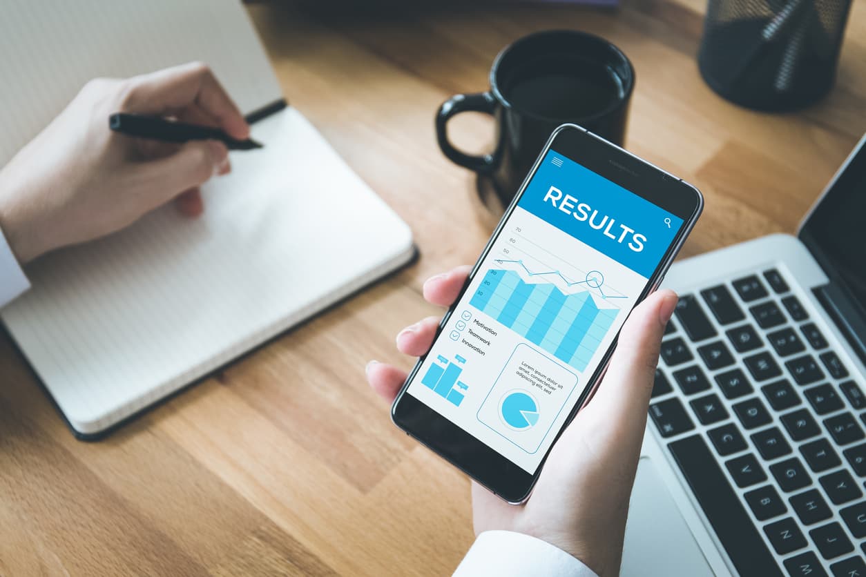 A businessperson holding a pen over a notebook and looking at a phone screen full of graphs that says “results”