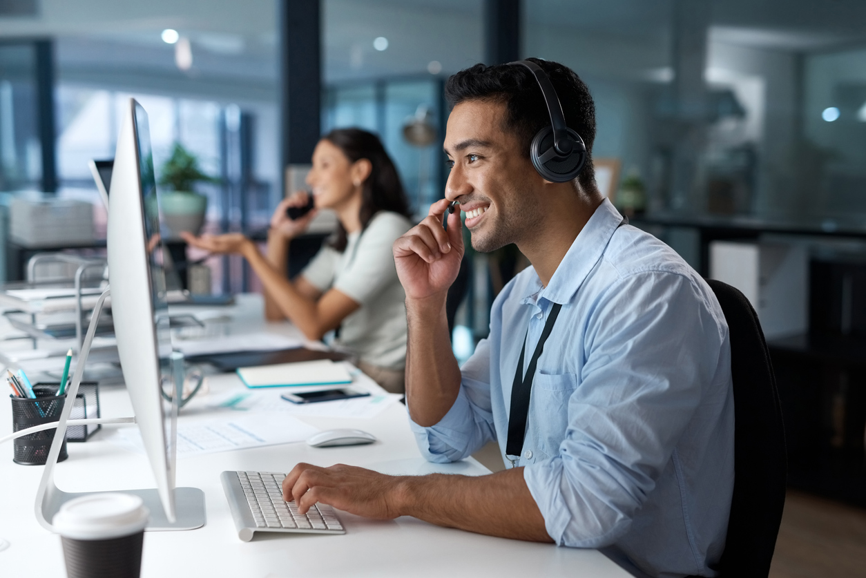 A young man sitting at a desk using a headset and computer for a sales call.